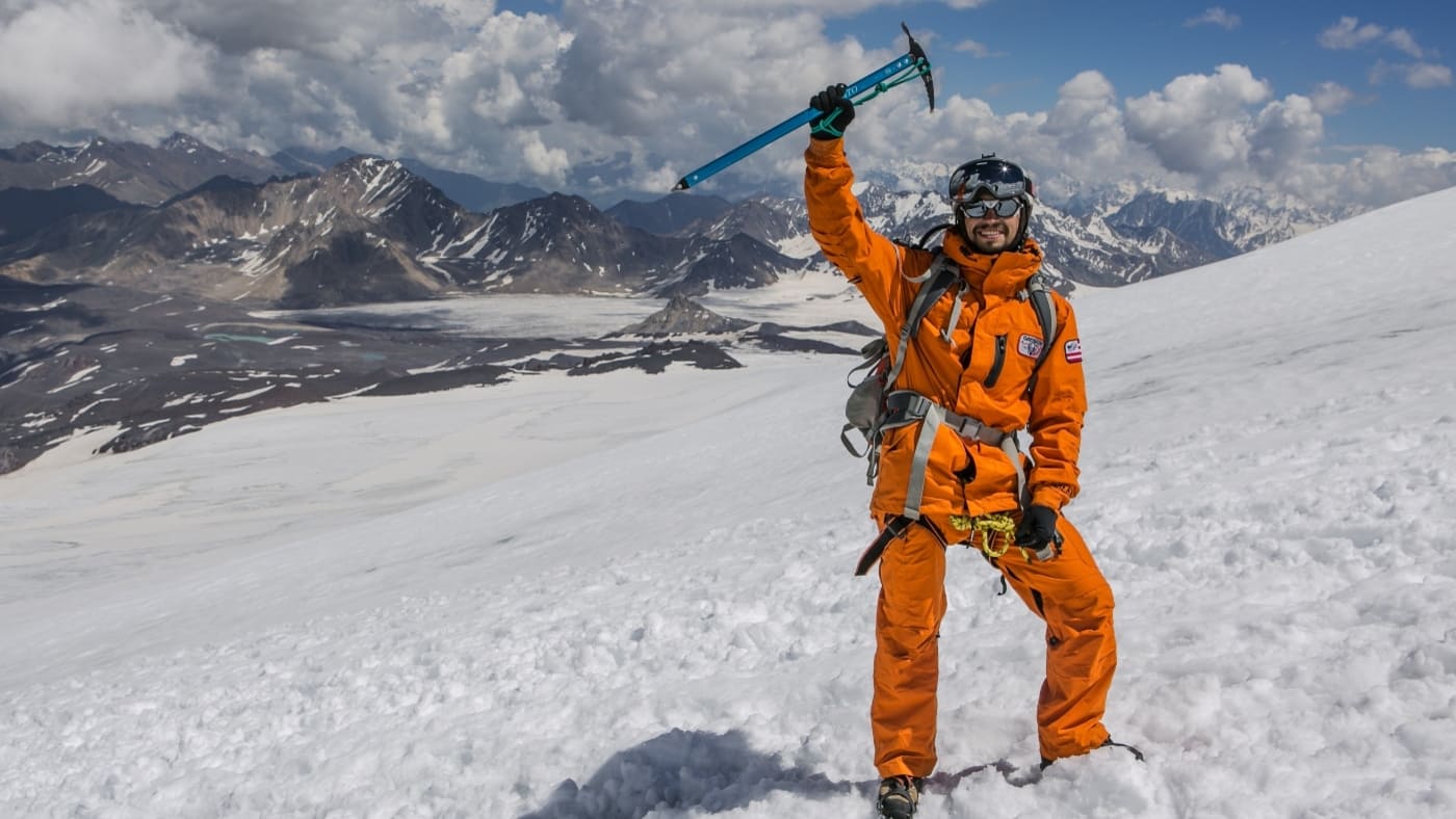 A climber triumphs on Elbrus, raising an ice axe against the stunning Caucasus back drop - B4Experience.