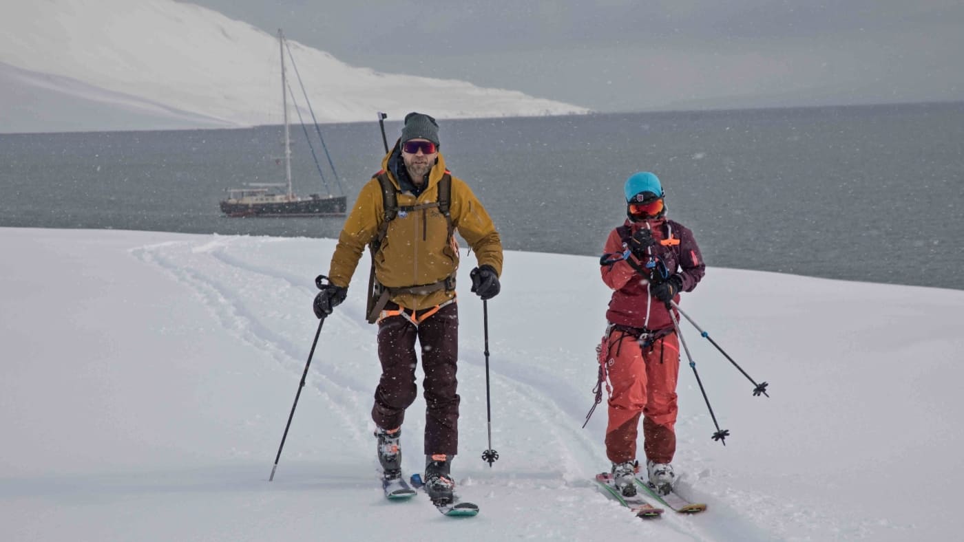 Couple of skiers, view of a sailboat at the bottom of the fjord.