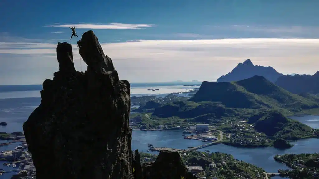 A climber. jumps between rock peaks in Svolvaer, with fjords below.