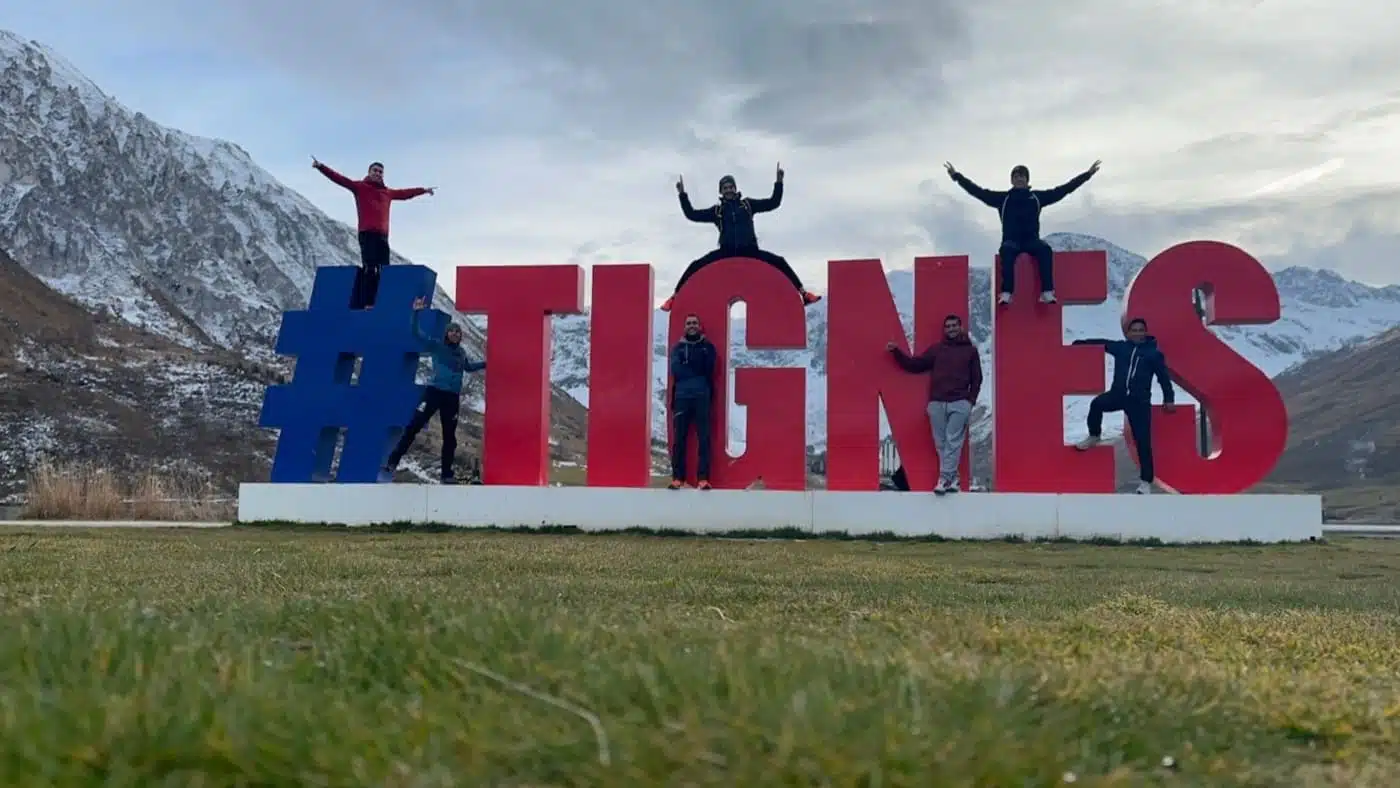 Group posing on a large "TIGNES" sign with mountains in the background with B4Experience