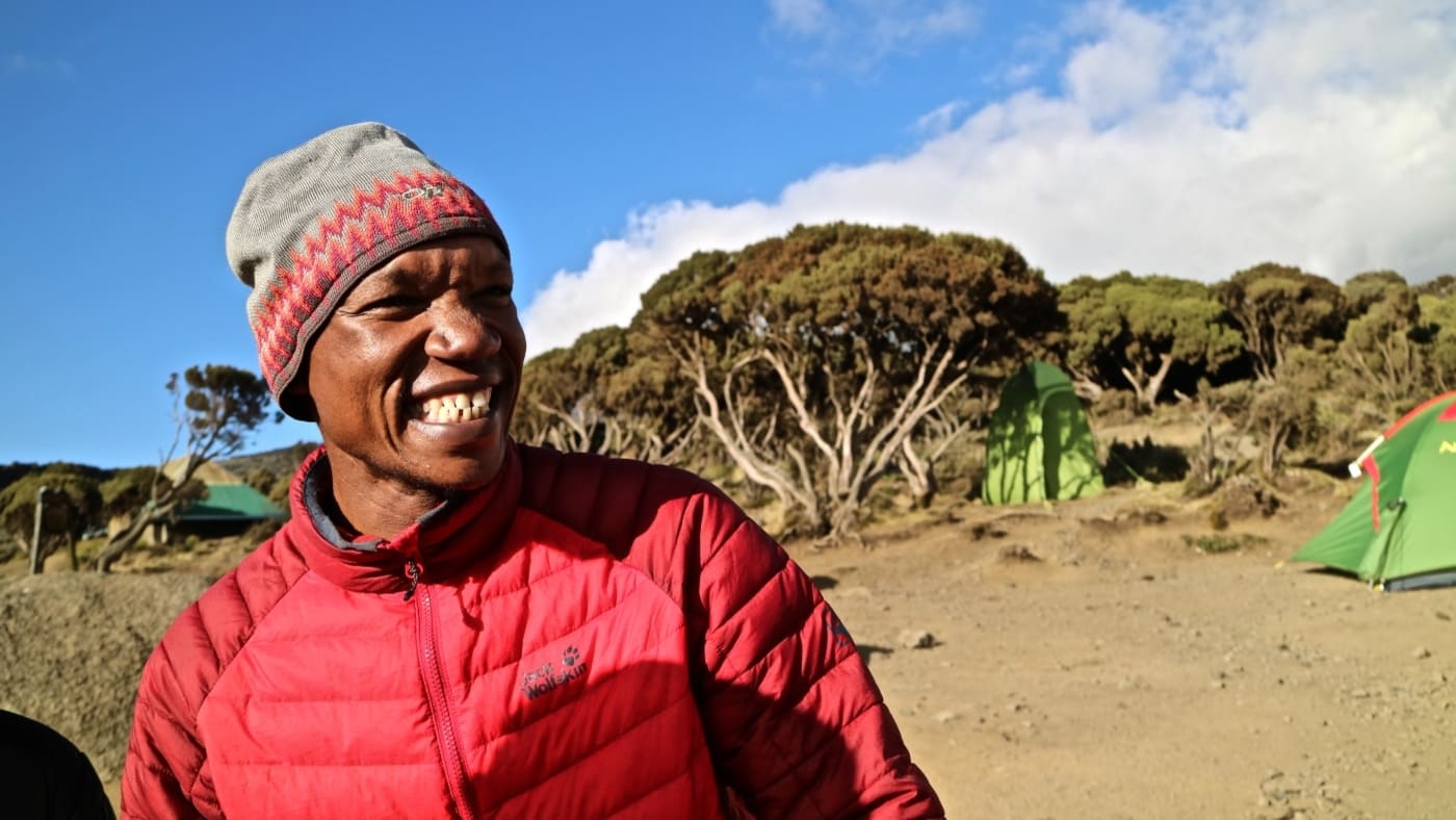 Smiling Kilimanjaro guide at a campsite with green tents in the background with B4Experience