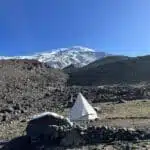 White tent in a rocky landscape with a snow-capped mountain under a clear blue sky - B4Experience