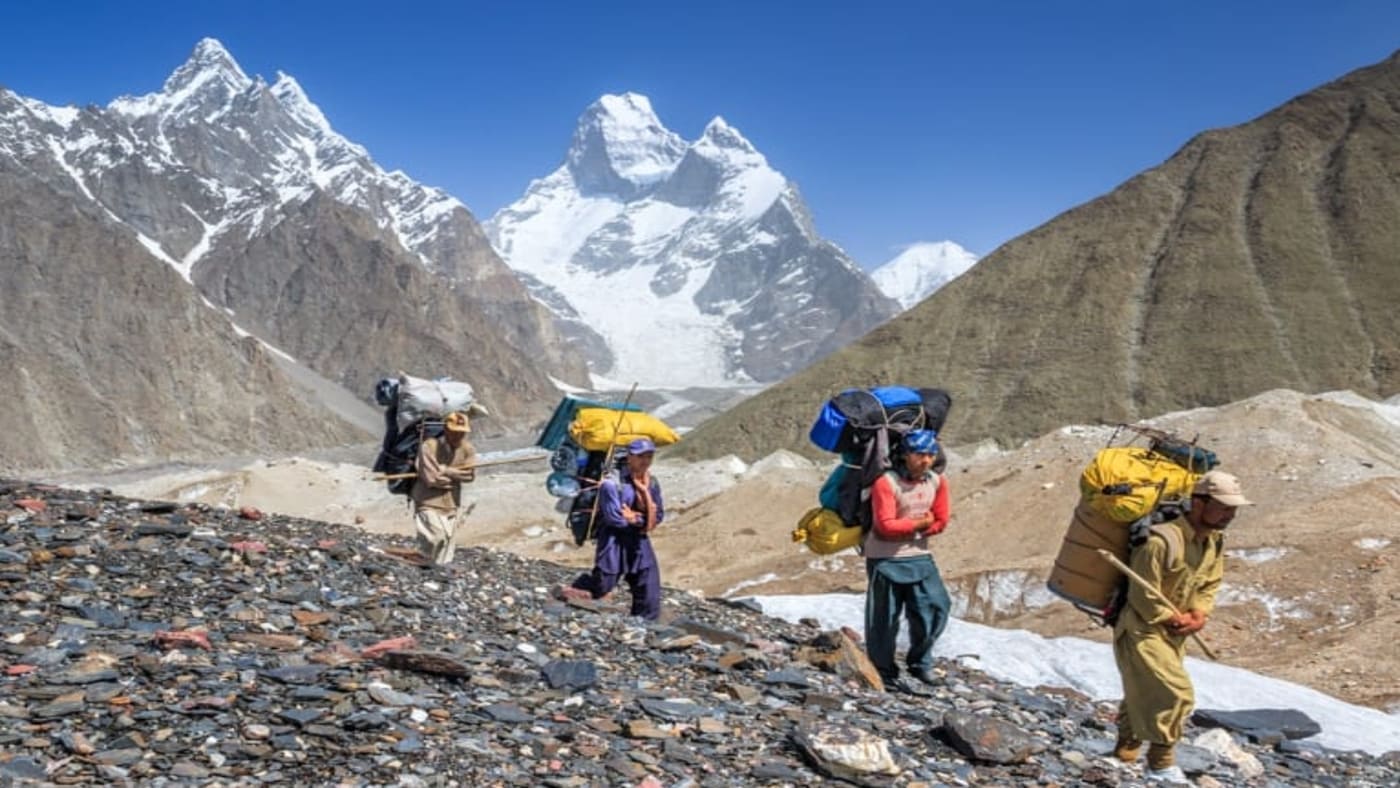 Group of trekkers and porters carrying heavy loads along a rugged trail with the K2 in the background - B4Experience